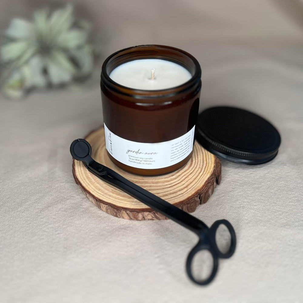 
                  
                    Sixteen-ounce Fern x Flow Garden Aura floral scented soy candle in an amber jar with black matte lid on the side right side and black matte wick trimmers diagonally across the front with a large air plant out-of-focus in the background.
                  
                