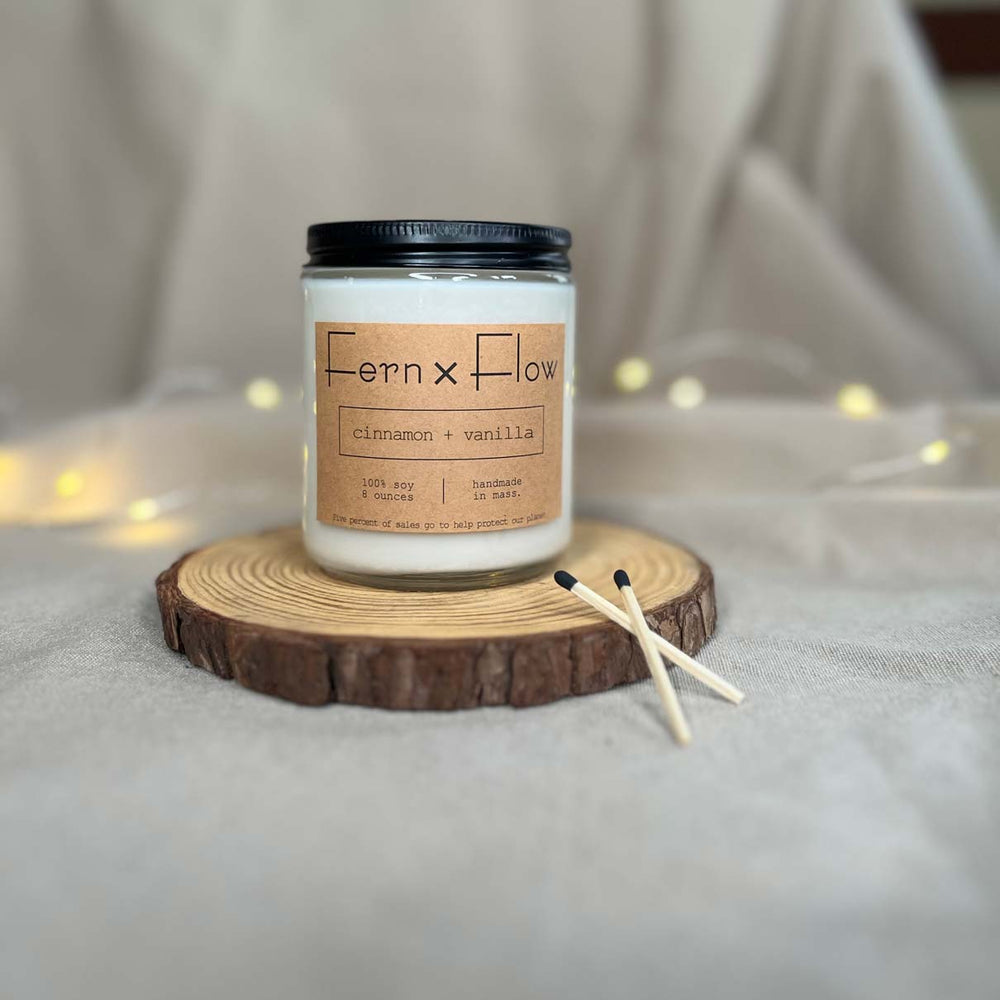 Fern x Flow eight-ounce Cinnamon + Vanilla scented soy candle on a rustic wooden riser with Christmas lights out-of-focus in the background.