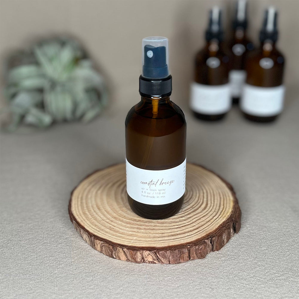 Fern x Flow natural and vegan 4oz Coastal Breeze scented Air + Linen Spray on a rustic wooden riser with other air and linen sprays out-of-focus in the background.
