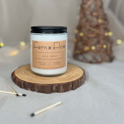 Fern x Flow eight-ounce Cozy Cabin scented soy candle on a rustic wooden riser with black-tipped safety matches in the foreground and a lighted, wicker tree in the background.