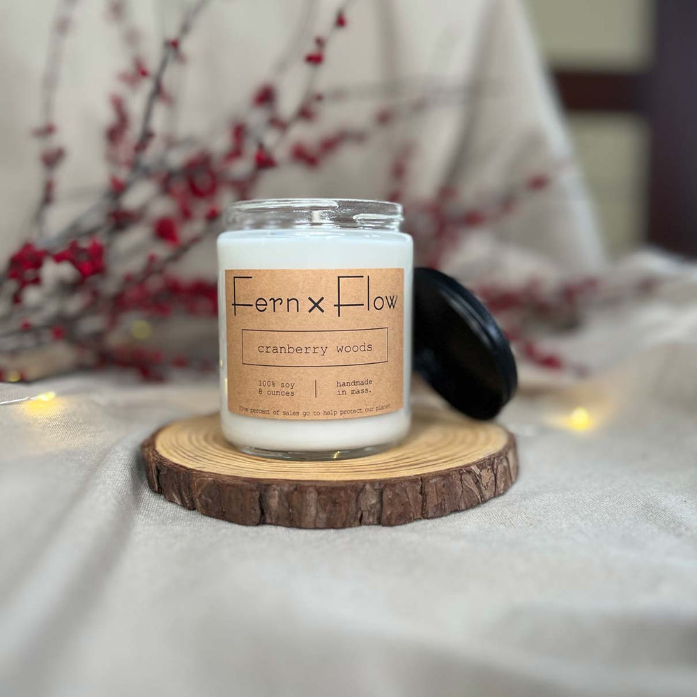 Fern x Flow eight-ounce Cranberry Woods scented soy candle on a rustic wooden riser with red berries and white lights out of focus in the background.