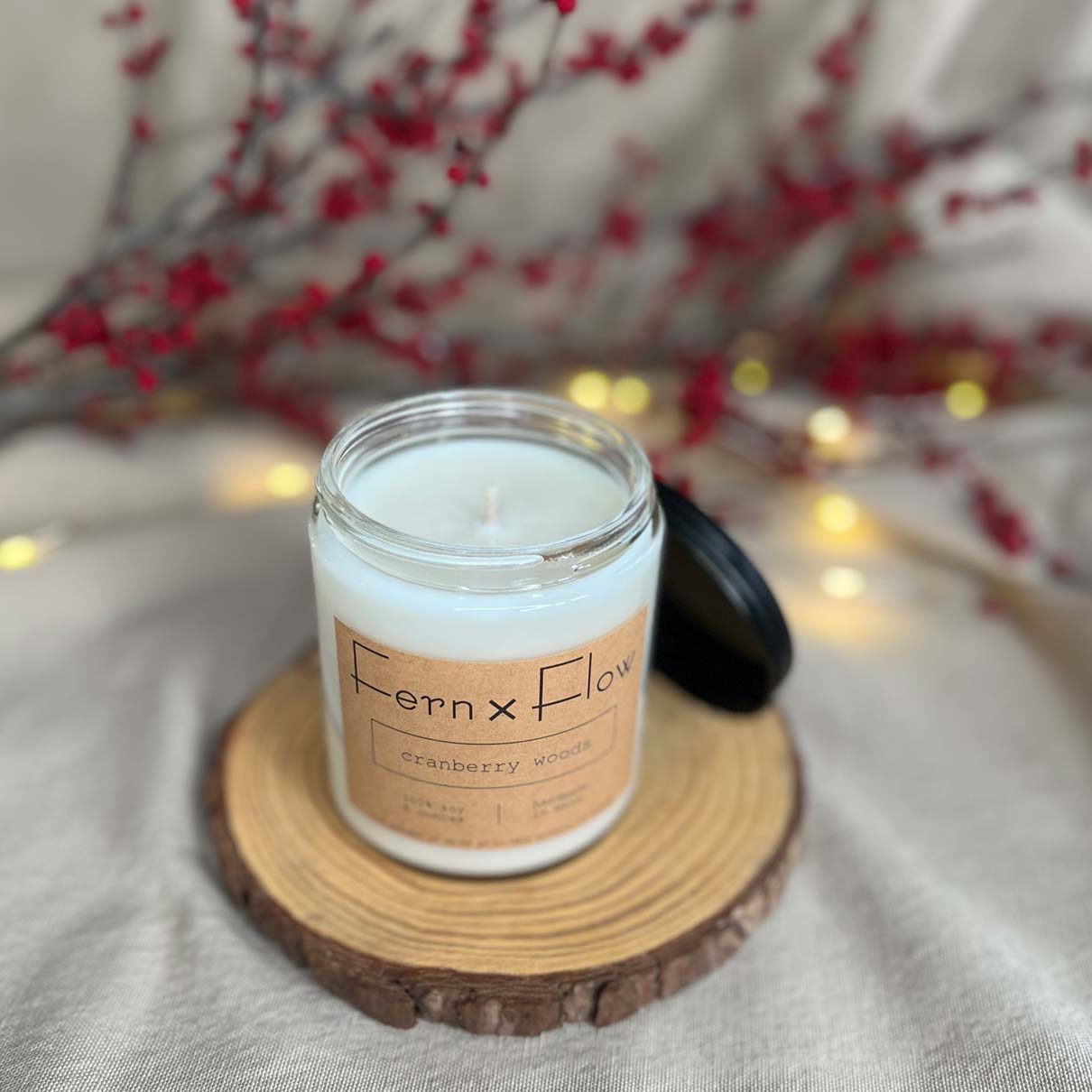 Fern x Flow Cranberry Woods scented soy candle on a rustic wooden riser with red berries and white lights out of focus in the background.