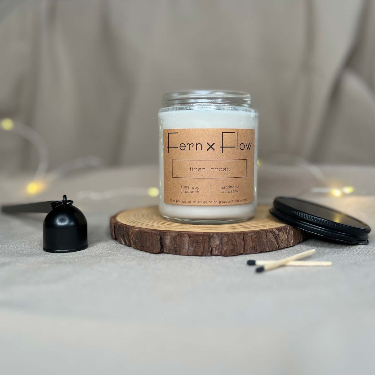 
                  
                    Fern x Flow eight-ounce First Frost scented soy candle on a rustic wooden riser with a black matte lid and candle snuffer on the sides.
                  
                
