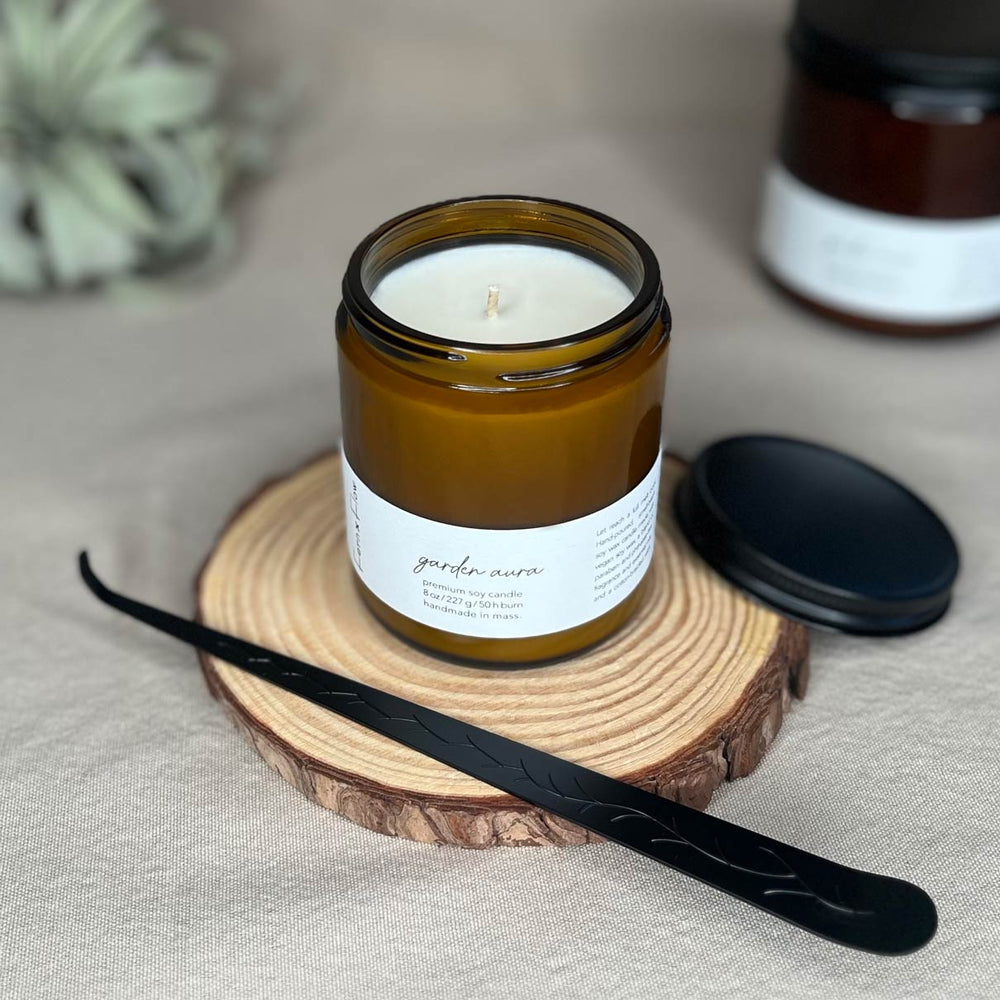 Eight-ounce Fern x Flow Garden Aura floral scented soy candle in an amber jar with black matte lid off to the side and black matte wick dipper diagonally across the front.