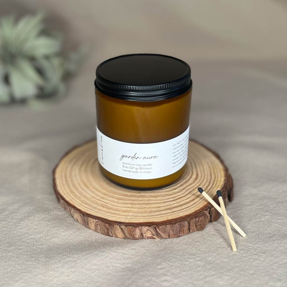 
                  
                    Eight-ounce Fern x Flow Garden Aura floral scented soy candle in an amber jar with black matte lid and two black-tipped safety matches crossed on the right side with a large air plant out-of-focus in the background.
                  
                