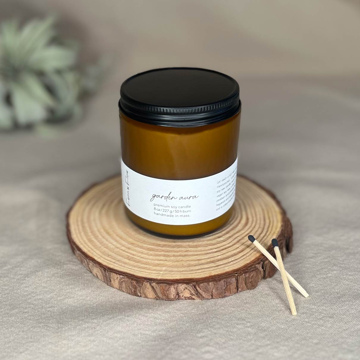 Eight-ounce Fern x Flow Garden Aura floral scented soy candle in an amber jar with black matte lid and two black-tipped safety matches crossed on the right side with a large air plant out-of-focus in the background.