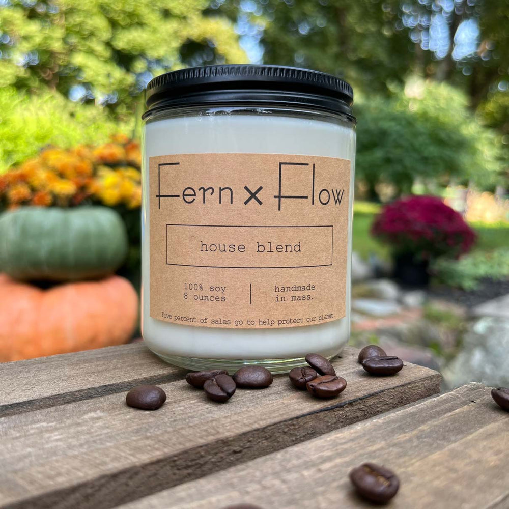 Fern x Flow House Blend coffee scented soy candle with coffee beans around the bottom of the glass jar, all atop a weathered, wooden crate with pumpkins and mums in the background.