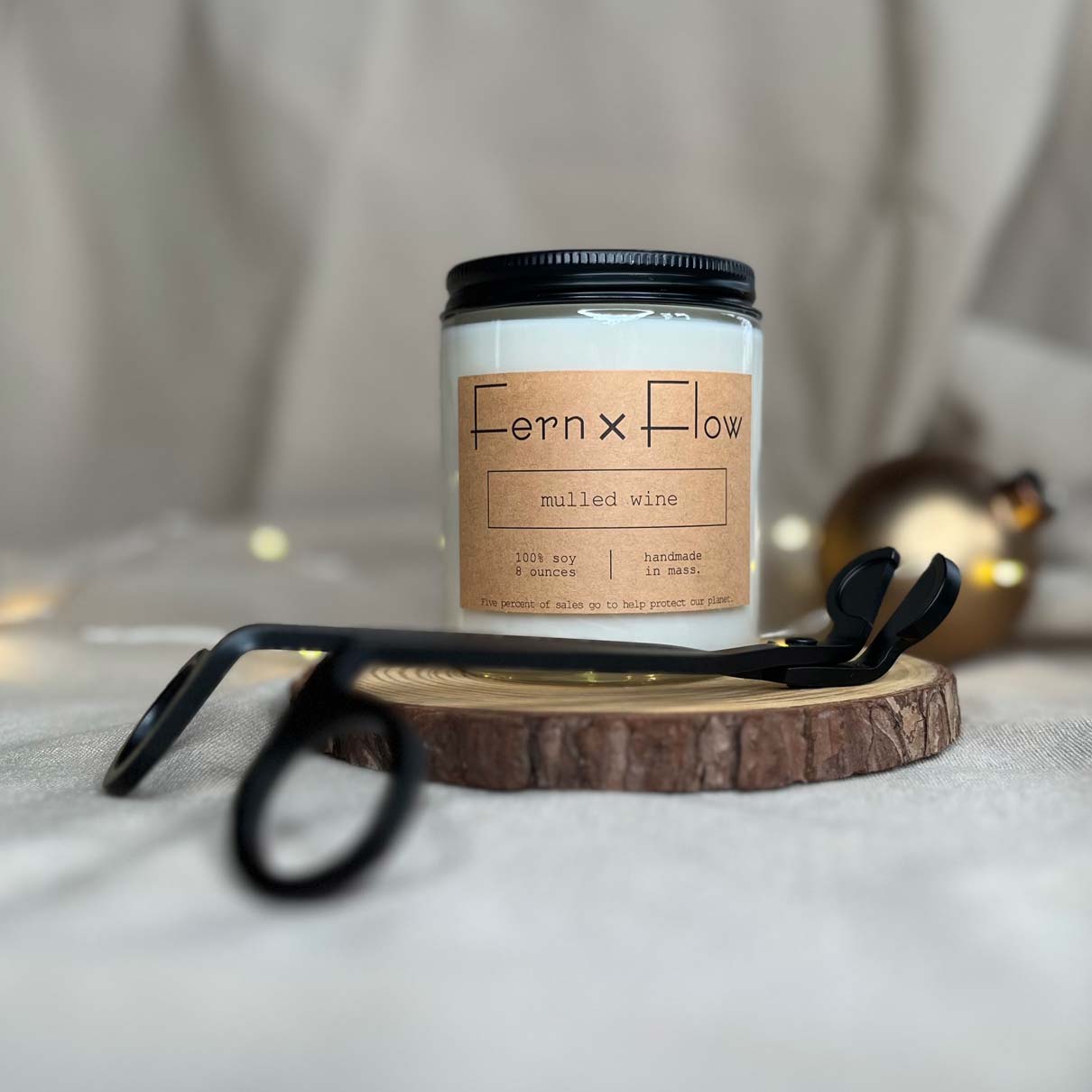 Fern x Flow eight-ounce Mulled Wine scented soy candle on a rustic wooden riser paired alongside matte black wick trimmers with white lights and golden ornament in the background.