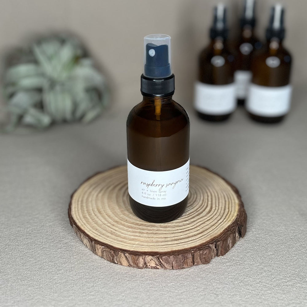 Fern x Flow natural and vegan 4oz Raspberry Sangia scented Air + Linen Spray on a rustic wooden riser with other air and linen sprays out-of-focus in the background.