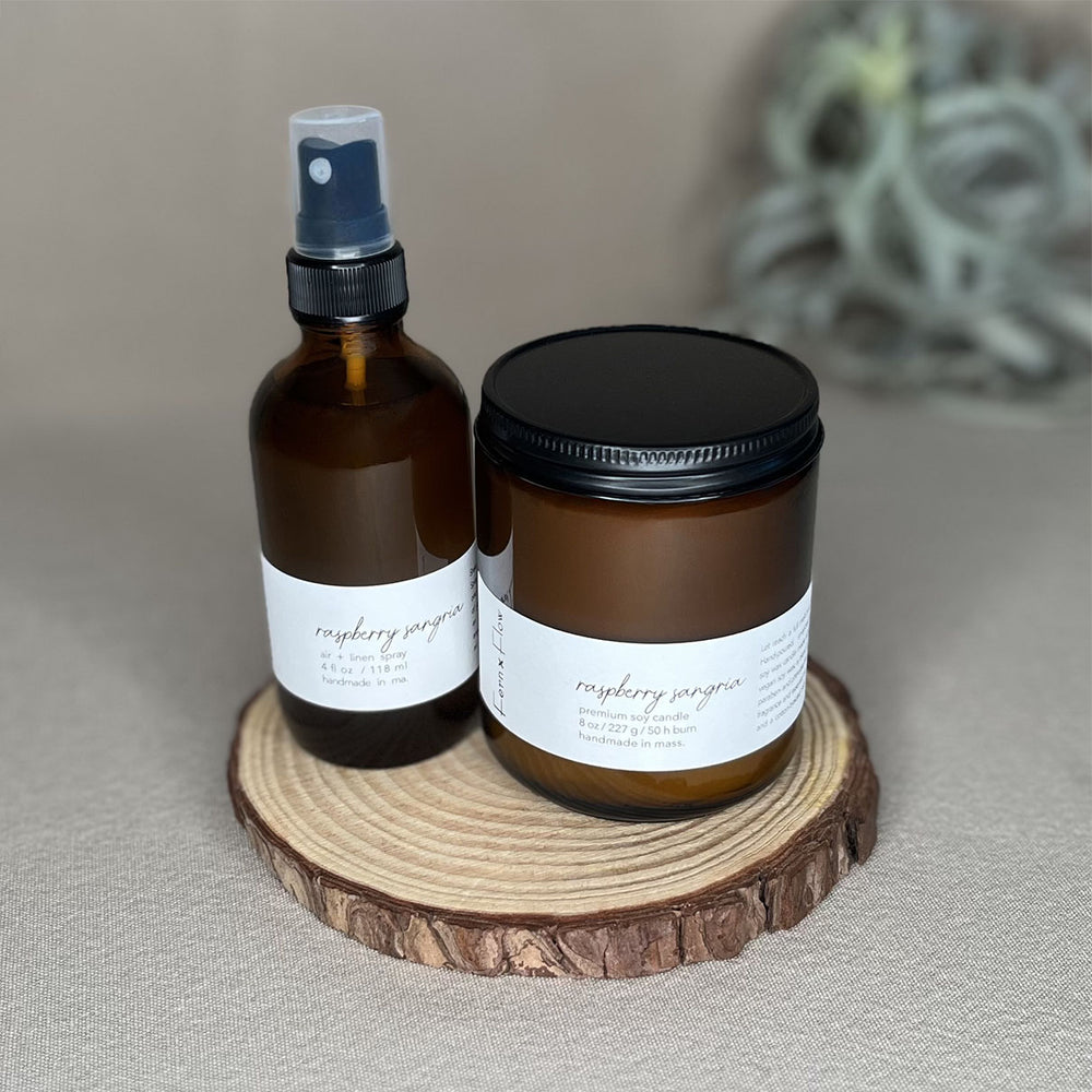 Fern x Flow Raspberry Sangria soy candle and air + linen spray fragrance bundle on a wooden rise in front of a large out-of-focus air plant in the background.