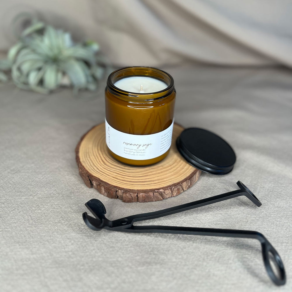 Eight-ounce Fern x Flow Rosemary Sage scented soy candle in an amber jar with the black matte lid and wick trimmers displayed against a cream-colored cloth background.