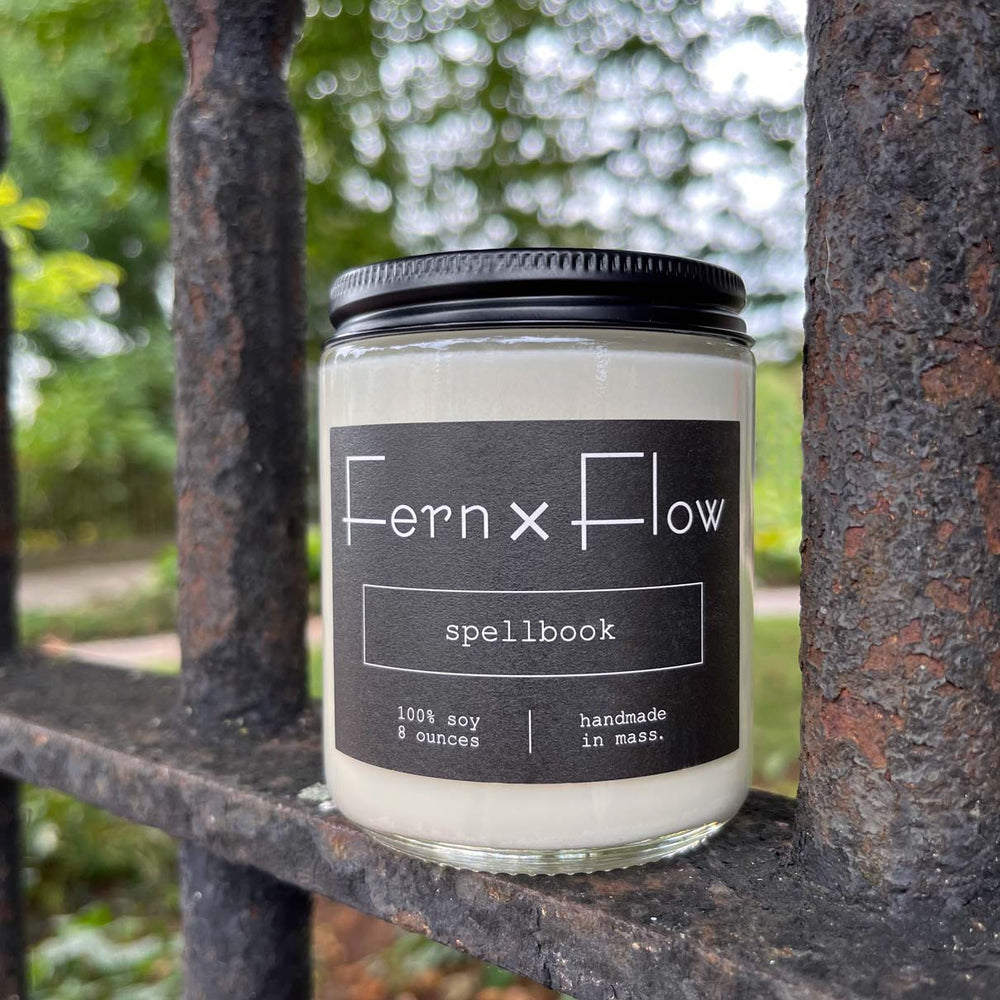 Fern x Flow Spellbook scented soy candle with a black and white label sitting on an old, black, wrought iron fence in Salem, MA.