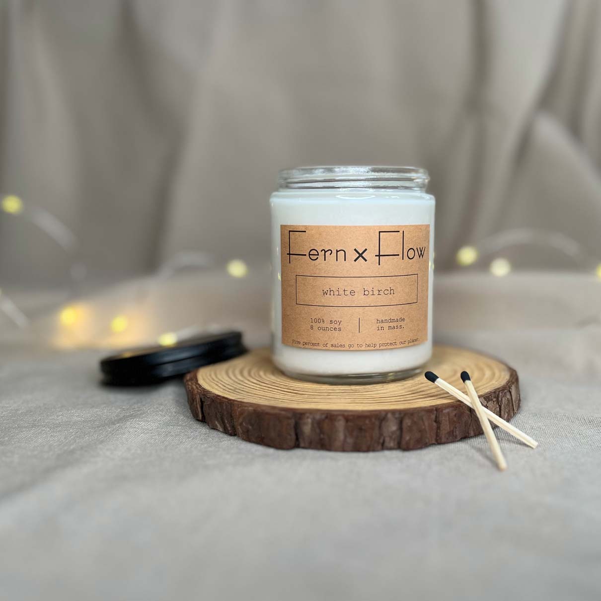 Fern x Flow eight-ounce White Birch scented holiday soy candle on a rustic wooden riser with black tipped safety matched in the foreground and white, Christmas lights in the background.