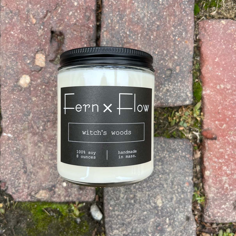 
                  
                    Fern x Flow Witch's Woods Halloween scented soy candle with black and white label against brick sidewalk in Salem, MA
                  
                