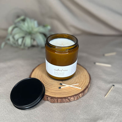 Eight-ounce Fern x Flow Woodland Acres scented soy candle on a rustic, wooden riser surrounded by black-tipped matches and a black matte lid with an air plant out-of-focus in the background.