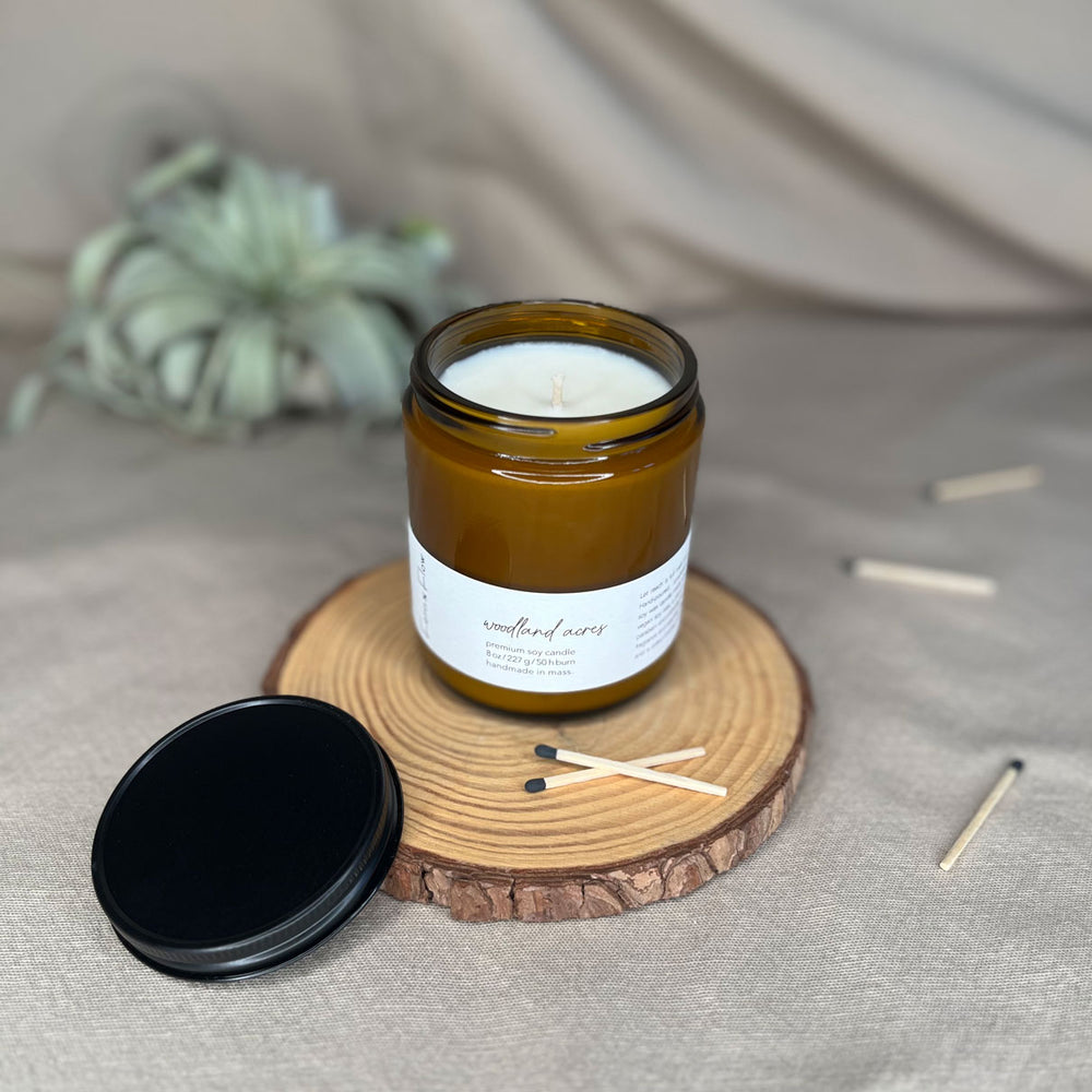 
                  
                    Eight-ounce Fern x Flow Woodland Acres scented soy candle on a rustic, wooden riser surrounded by black-tipped matches and a black matte lid with an air plant out-of-focus in the background.
                  
                