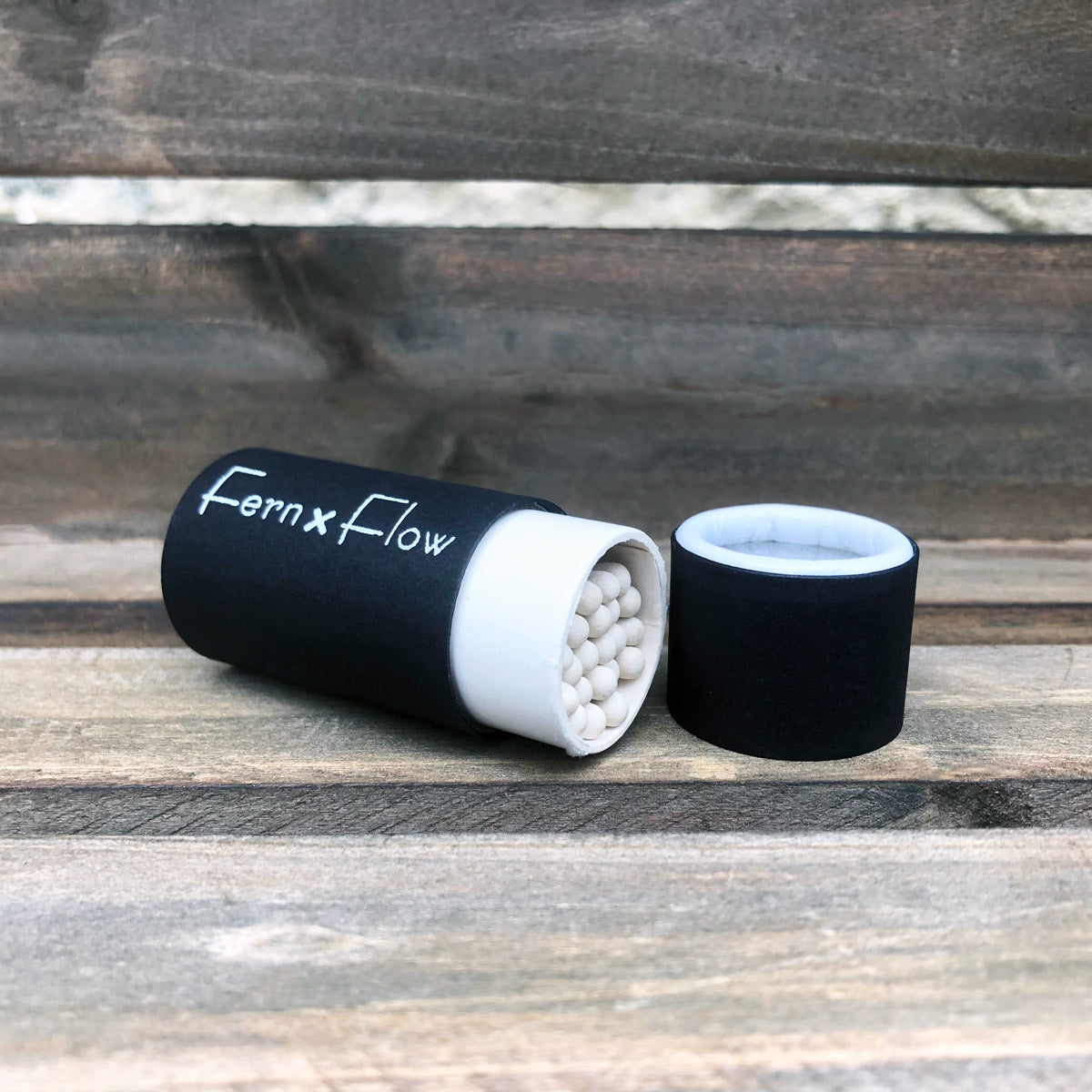 
                  
                    Fern x Flow, matte black, safety barrel match stick bottle lying on it's side at a 45-degree angle with white-tipped matches showing and the cap to the right, against a weathered, wooden background.
                  
                