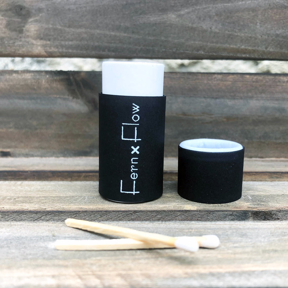 Fern x Flow, matte black, safety barrel match stick bottle opened with two white-tipped safety matches crossing in the foreground, with a weathered, wooden background.