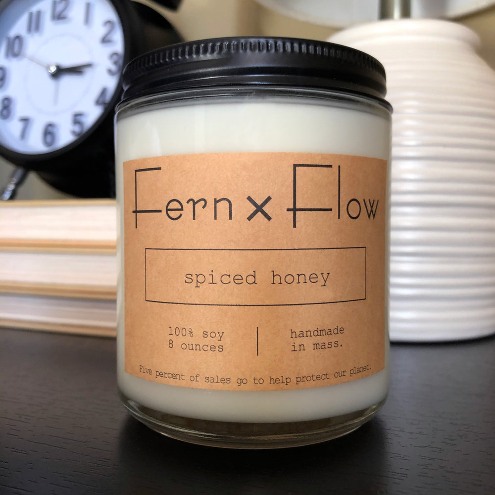 
                  
                    Eight-ounce Fern x Flow Spiced Honey scented soy candle on a black table, in front of a black alarm clock, on a stack of books and a white, modern lamp.
                  
                